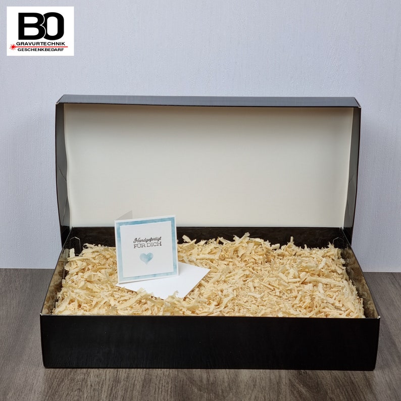 May include: A black gift box with a white interior, filled with wood shavings. A small card with the text "Handgefertigt F&uuml;r Dich" and a blue heart is placed inside the box. The box is open, revealing its contents.