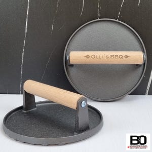 Könnte beinhalten: Zwei Burgerpressen aus Gusseisen mit Holzgriffen. Eine Presse steht aufrecht, die andere liegt flach. Die Holzgriffe sind mit dem Text "Olli's BBQ" graviert. Die Pressen sind dunkelgrau und rund.