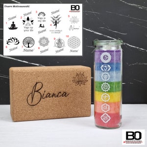 Könnte beinhalten: Ein Set an Yoga-Geschenken, bestehend aus einem Kork-Yogablock mit dem eingravierten Namen "Bianca", einem Glasgefäß mit bunten Stoffschichten und weißen Symbolen sowie einem Blatt Papier mit zehn verschiedenen Yoga-Motiven.