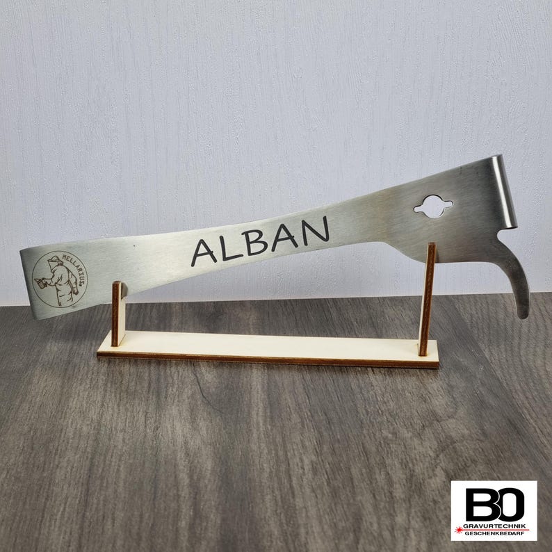 May include: A stainless steel beekeeping tool with the name "ALBAN" engraved on it. The tool is displayed on a wooden stand. The tool has a hook, a circular hole, and a logo with a person holding a tool.