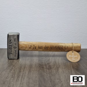 Peut inclure: Un marteau personnalisé avec un manche en bois. La tête du marteau est gravée du nom "TOBIAS" et du texte "Original seit 1995". Le manche est gravé du texte "Du bist der HAMMER" et de deux cœurs. Une étiquette brune avec le texte "Handmade with love" est attachée au manche.