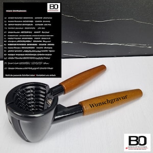 Könnte beinhalten: Ein schwarzer Nussknacker mit Holzgriffen, mit dem eingravierten Wort "Wunschgravur". Das Bild enthält eine Liste von Schriftoptionen und ein Logo in der oberen rechten Ecke. Der Nussknacker ist zum Knacken von Nüssen bestimmt.