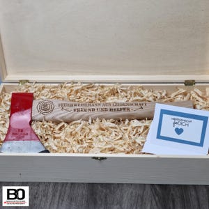 Peut inclure: Une boîte en bois contenant une hache rouge et argent. Le manche de la hache est gravé du texte "Feuerwehrmann aus Leidenschaft - Freund und Helfer" et d'un logo de pompier. Une petite carte avec le texte "Handgemacht für Dich" est également dans la boîte.