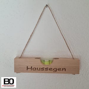 Könnte beinhalten: Ein Holz-Wasserwaage mit einer grünen Blase und dem Text "Hausseegen" eingraviert. Die Wasserwaage hängt an einer Schnur.
