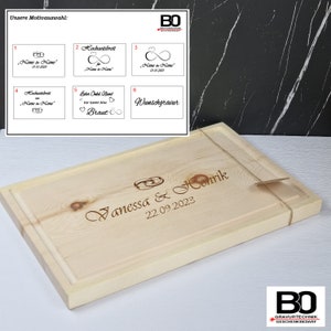 Op de afbeelding: Een houten snijplank met een gepersonaliseerde inscriptie die "Vanessa & Henrik 22.09.2023" luidt. De inscriptie is in een cursief lettertype en bevat een ontwerp van twee in elkaar verweven ringen. De plank is lichtbruin en heeft een verhoogde rand.