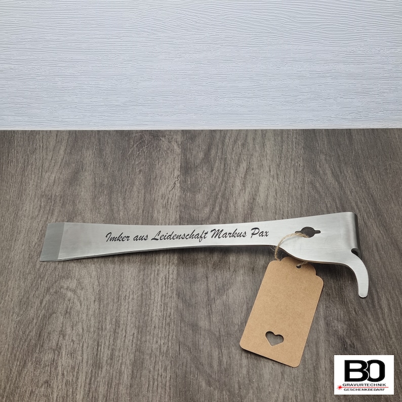 May include: A stainless steel beekeeping hive tool with the engraved text "Imker aus Leidenschaft Markus Pax". The tool has a flat end, a hook, and a small hole. A brown tag with a heart cutout hangs from the tool.