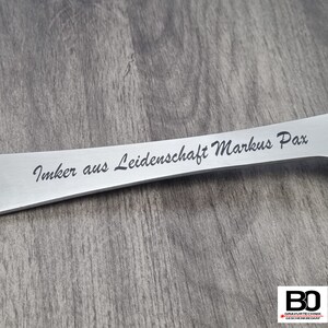 May include: A stainless steel beekeeping tool with the engraved text "Imker aus Leidenschaft Markus Pax". The tool has a flat, elongated design with a slightly curved shape. The background is a wooden surface.