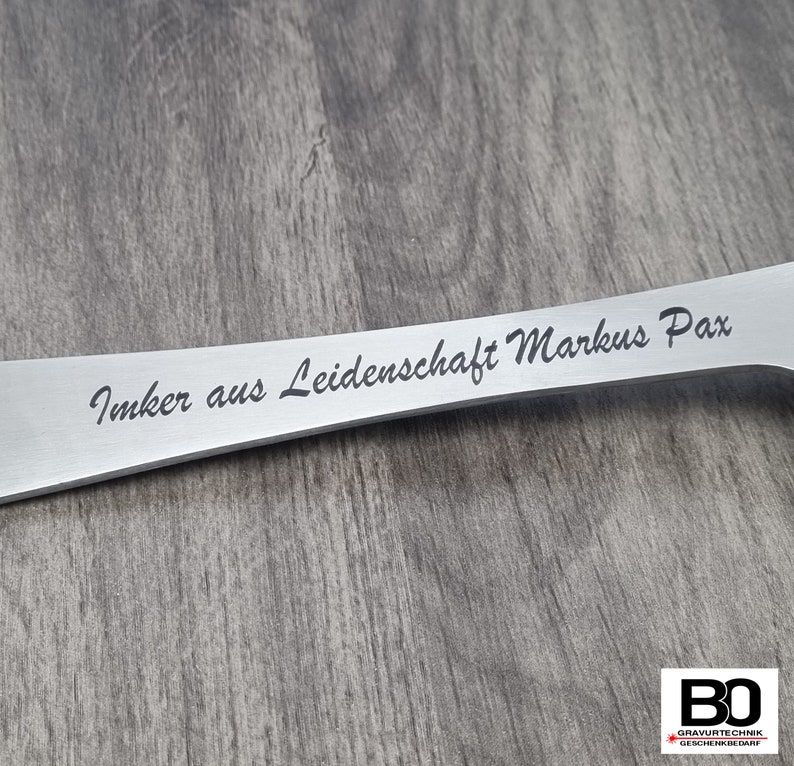 May include: A stainless steel beekeeping tool with the engraved text "Imker aus Leidenschaft Markus Pax". The tool has a flat, elongated design with a slightly curved shape. The background is a wooden surface.