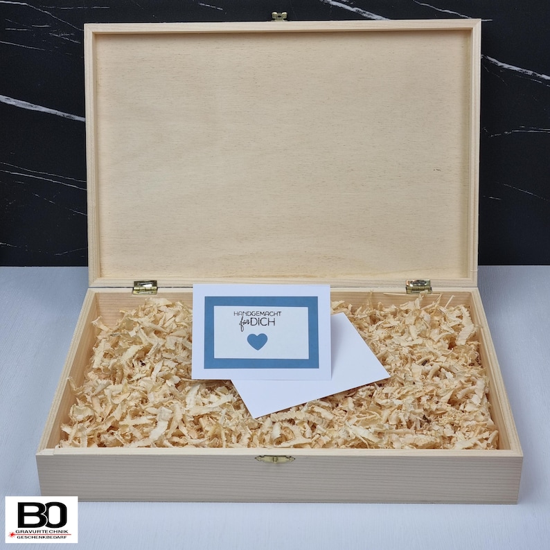 May include: Open wooden box with a hinged lid, filled with wood shavings. A card with the text "Handmade for you" and a blue heart is inside the box, along with a white envelope. The box is made of light-colored wood.