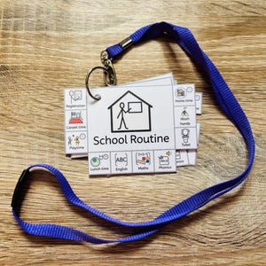 May include: A set of white rectangular cards featuring black line drawings and text for a school routine, attached to a silver metal ring and a blue fabric lanyard. The cards display icons for activities like registration, carpet time, and lunch time.