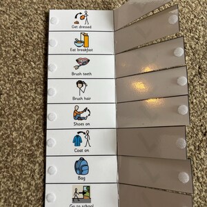 May include: A visual timetable for a morning routine, with illustrated steps including "Get dressed," "Eat breakfast," and "Go to school." The schedule is made of white card with a series of attached, grey, laminated flaps. Each step has an image and text.