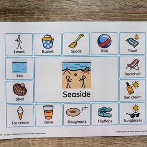 May include: A white card featuring beach-related illustrations such as a bucket, ball, and sunglasses. The word "Seaside" is in the center. The card is on a wooden surface.