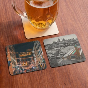 May include: Two square coasters with different cityscapes. One coaster features a nighttime view of a city street with string lights, while the other shows a daytime aerial view of a city skyline with a large building in the foreground.