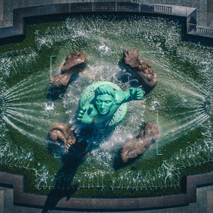 May include: An aerial view of a fountain with a green statue of a person reaching out with their arms. The statue is surrounded by four brown statues of animals, and the fountain is filled with water.