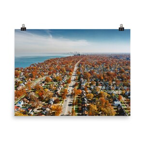 May include: An aerial view of a suburban neighborhood in the fall. The houses are surrounded by trees with colorful autumn leaves. A long road runs through the center of the neighborhood, leading to a city skyline in the distance.