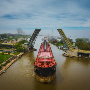 May include: A large red and black cargo ship passes through a raised drawbridge. The bridge is made of steel and has a yellow stripe down the center. The ship is in the middle of the channel, with water on either side. The sky is cloudy and there are trees and buildings in the background.