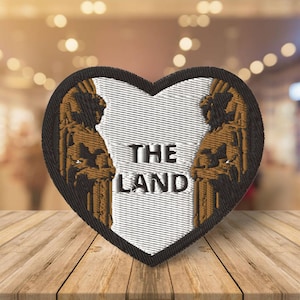 May include: A heart-shaped embroidered patch with a white background and black outline. The patch features two brown figures with wings on either side of the text "THE LAND".