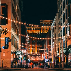 May include: A night view of a city street lined with brick buildings and illuminated with string lights. The street is empty except for a few people walking in the distance. The buildings are decorated with signs and lights, creating a festive atmosphere.