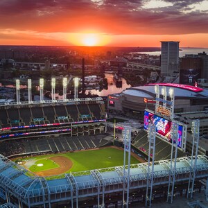 May include: An aerial view of a baseball stadium at sunset with a scoreboard displaying "Indians" and "World Series". The stadium is surrounded by a city skyline and a river.