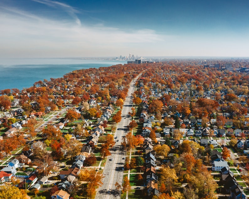 May include: Aerial view of a suburban neighborhood with houses and trees in various shades of autumn colors. A long road runs through the center of the image, leading to a city skyline in the distance.