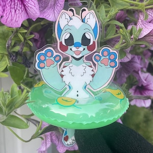 Puede incluir: Un animal de dibujos animados azul claro y blanco con mejillas rojas está sentado en un flotador de piscina inflable verde. El animal tiene ojos grandes y una expresión feliz. El flotador de piscina tiene acentos amarillos.