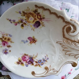 Könnte beinhalten: Eine weiße Porzellanschale mit einem Blumendesign in Rosa-, Gelb- und Blautönen. Die Schale hat einen gewellten Rand und einen goldbemalten Griff.