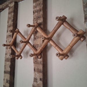 May include: A vintage wooden accordion-style coat rack with multiple pegs. The rack is mounted on a wall with exposed wooden beams. The coat rack is made of light brown wood and has a geometric design. It is a functional and decorative item.