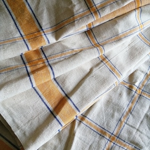 Pode incluir: Uma toalha de cozinha de linho branco com listras azuis e amarelas. A toalha está dobrada ao meio e as listras são visíveis em ambos os lados.