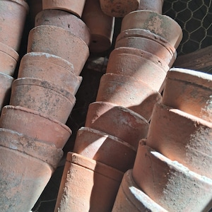 Op de afbeelding: Een stapel terracotta bloempotten, sommige zijn gestapeld in groepen van drie of vier, andere in groepen van twee. De potten zijn allemaal van verschillende grootte en hebben een rustieke, aardse uitstraling.