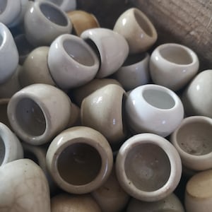 May include: A close-up of a collection of small, off-white ceramic bowls. The bowls have a rounded shape and open tops. The bowls are clustered together, showing variations in colour and texture.