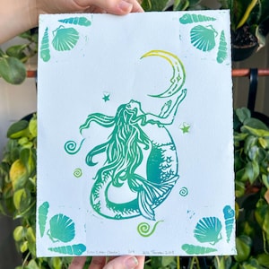 May include: A green and blue linocut print of a mermaid holding a crescent moon. The mermaid has long flowing hair and a fishtail. The print is surrounded by seashell and spiral designs. The print is held by a person in front of a background of green plants.