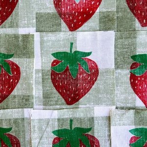 May include: A close-up of a fabric featuring a repeating pattern of red strawberries with green leaves set against a green and white checked backdrop. The strawberries have white seeds.