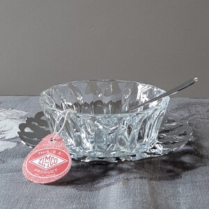 May include: A clear glass bowl with a decorative pattern sits on a silver metal base. A small silver spoon rests inside the bowl. A red and white tag with the text "This is a Zimco Product" is attached to the bowl.