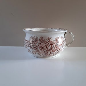 Puede incluir: Orinal antiguo de cerámica blanca con un diseño floral de rosas marrones. El orinal tiene un asa y un borde ancho. El diseño floral se repite en el exterior. Una pieza decorativa vintage.