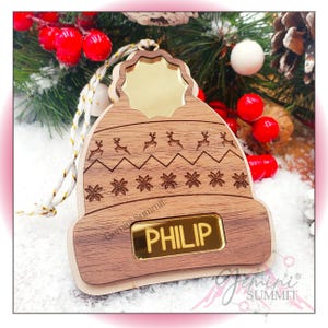 May include: Wooden Christmas ornament shaped like a brown knit hat with a gold star on top. The hat has a white snowflake pattern and a gold name tag that says "PHILIP".