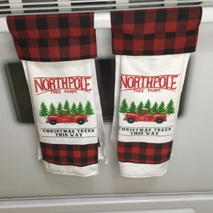 May include: Two white kitchen towels with a red and black buffalo plaid top and bottom. The towels have a red graphic of a truck carrying Christmas trees with the text "NORTHPOLE TREE FARM" and "CHRISTMAS TREES THIS WAY."