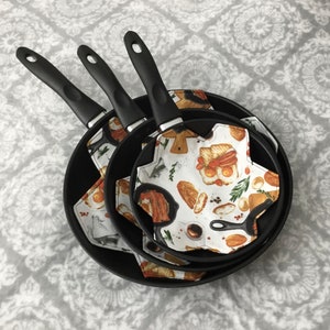 May include: A set of three black non-stick frying pans with black handles. Each pan contains a patterned trivet with a breakfast food design. The trivets feature images of eggs, bacon, toast, and coffee.
