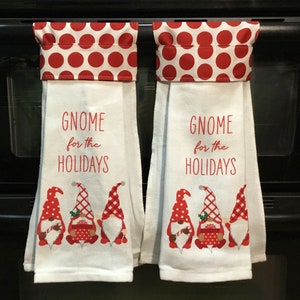 May include: Two white kitchen towels with red polka dot tops and the text "Gnome for the Holidays." Each towel features a row of gnome illustrations in red and white. The towels are hanging on a black oven.