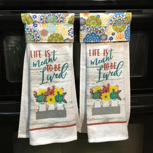 May include: Two white kitchen towels with the text "LIFE is meant TO BE Lived" and a floral design. The towels have a decorative top with a colorful floral pattern. The towels are hanging on a black oven.