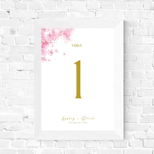 May include: White framed table number card with a pink watercolor floral design in the top left corner. The number "1" is in gold and the text "TABLE" is in black. The text "Abby & David" is in black cursive script with the date "September 18, 2022" in black below.