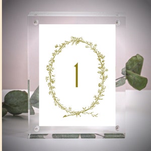 May include: A clear acrylic table number holder with a white card inside. The card features a gold floral wreath with the number "1" in the center.