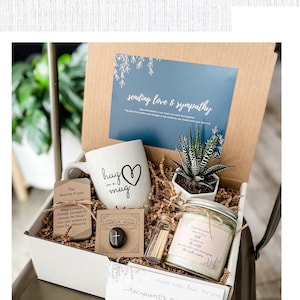 K&ouml;nnte beinhalten: Eine wei&szlig;e Geschenkbox gef&uuml;llt mit Mitgef&uuml;hlsgaben, darunter eine Tasse mit der Aufschrift "Hug is in the mug", eine kleine Sukkulente, eine Kerze, ein Gebetstein und eine handgeschriebene Karte. Die Box ist mit einem Etikett verziert, auf dem "Sending love & sympathy" steht.