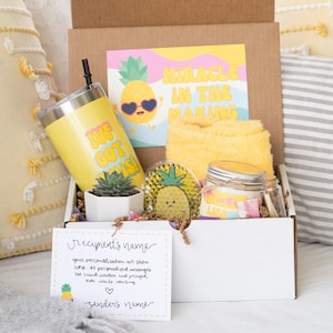 May include: A white gift box filled with a variety of items, including a yellow tumbler with the text "Live, Cot, Crush", a yellow towel with the text "Miracle in the Making", a small potted succulent, a pineapple-shaped candle, and a card with the text "recipient's name" and "sender's name".