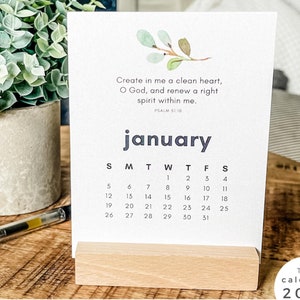 Puede incluir: Calendario de enero de 2025 sobre un soporte de madera. El calendario presenta una cita y un diseño minimalista. Un bolígrafo y una planta en maceta están sobre la superficie de madera.