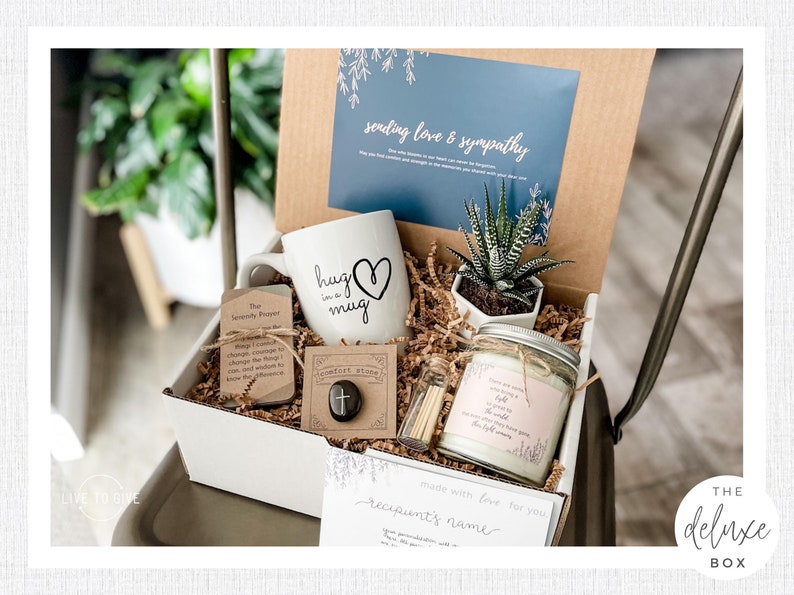 K&ouml;nnte beinhalten: Eine wei&szlig;e Geschenkbox gef&uuml;llt mit Artikeln f&uuml;r einen Beileids-Geschenk-Korb. Die Box enth&auml;lt eine wei&szlig;e Tasse mit dem Text "hug in a mug" und einem Herzen, eine kleine Sukkulente in einem wei&szlig;en Topf, eine Kerze mit dem Text "Sending love and light to a world that needs it", einen kleinen Troststein und eine Karte mit dem Text "made with love for you, recipient's name".