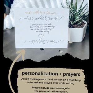 May include: A white card with the text "made with love for you..." and a space for the recipient's name. The card also includes a space for the sender's name and a heart symbol. The text "Your personalization will show here. All personalized messages are handwritten and prayed over while writing." is also included on the card. The card is sitting on a white surface with a small potted plant in the background.