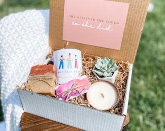 May include: A white gift box filled with a variety of items, including a mug with a colorful illustration of women, a bar of soap, a small succulent plant in a white pot, a candle, and a card with the text "The future is female". The box is sitting on a wooden table. A pink card with the text "She believed she could so she did." is visible in the background.