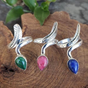 Può includere: Tre anelli d'argento con design a piuma e gemme a goccia. Un anello presenta una gemma verde, un altro una gemma rosa e il terzo una gemma blu. Gli anelli sono esposti su una superficie di legno.