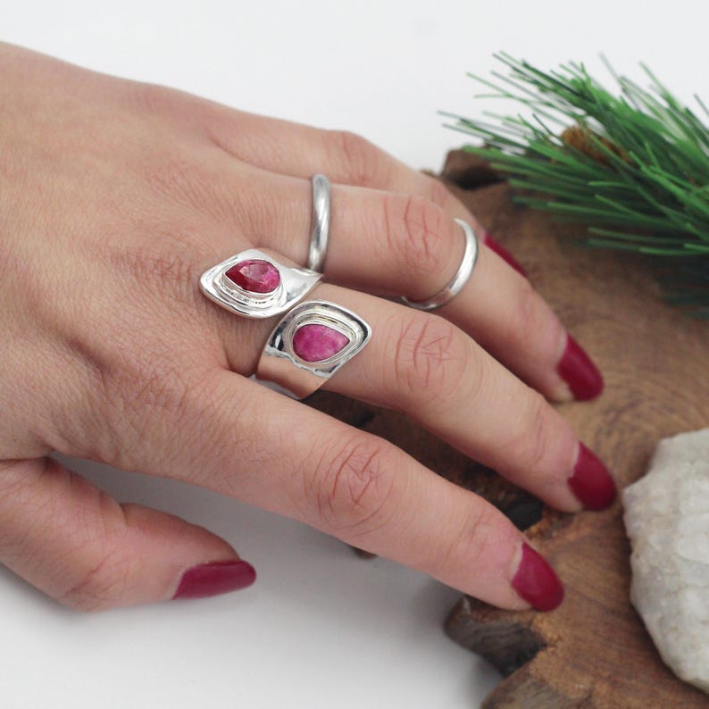 Może przedstawiać: Dłoń prezentuje trzy srebrne pierścionki. Dwa pierścionki mają kształt łzy, jasnoczerwony kamień szlachetny osadzony w srebrnej oprawie. Trzeci pierścionek to prosta srebrna obrączka. Dłoń ma czerwony lakier do paznokci.