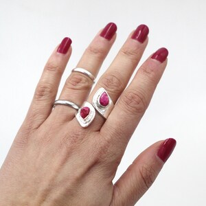 Może przedstawiać: Dłoń z trzema srebrnymi pierścionkami, w tym dwoma z dużymi, czerwonymi kamieniami w kształcie łezki. Pierścionki są na białym tle. Paznokcie pomalowane są na głęboki czerwony kolor.